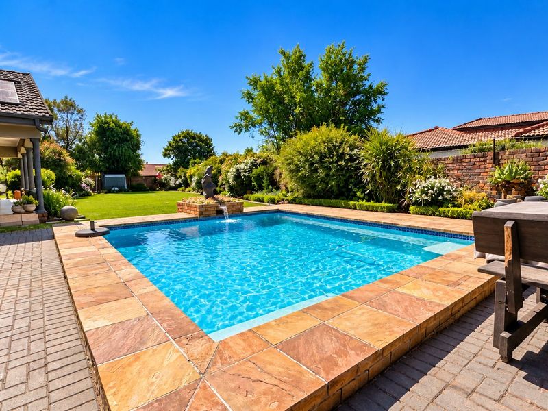 A well-maintained outdoor pool area featuring stone coping, a water feature, and a paved patio, set within a landscaped garden with a brick boundary wall.