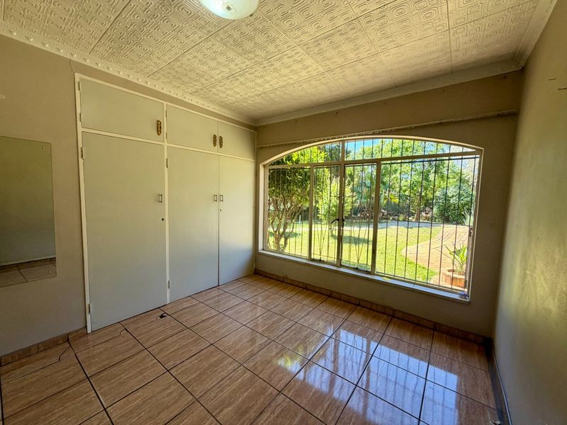 A vacant bedroom featuring tiled flooring, built-in cupboards, and an arched window with security bars overlooking the garden.