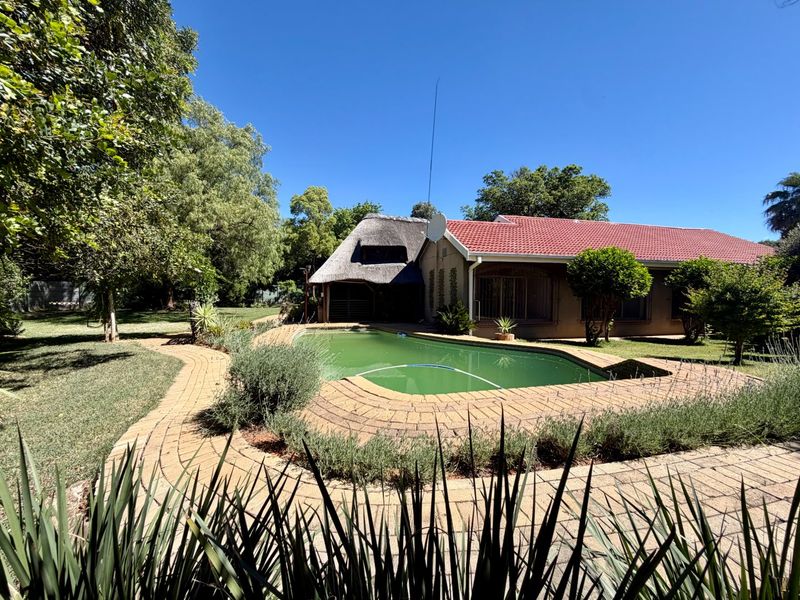 Rear garden view featuring a swimming pool with paved surround, mature landscaping, and a combination of thatched and tiled roof structures.