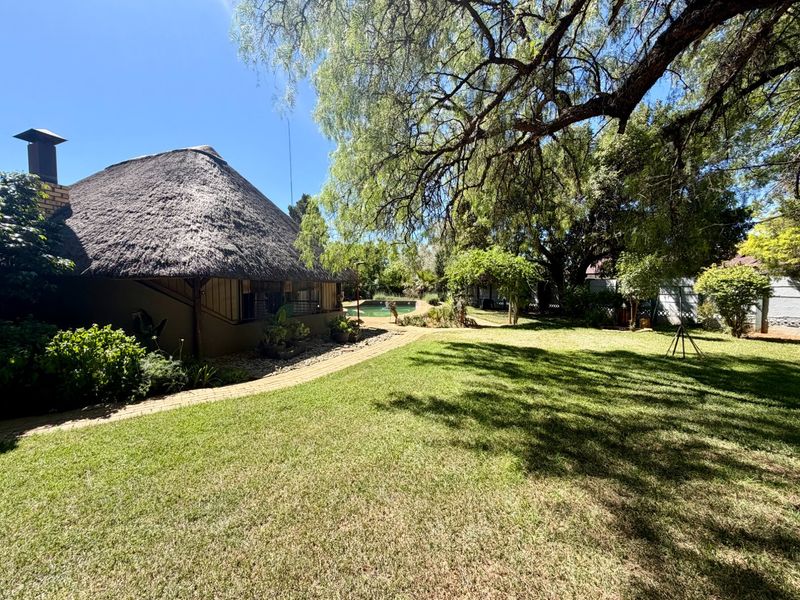 Spacious rear garden featuring a well-maintained lawn, paved pathway, swimming pool, and a house with a traditional thatch roof.