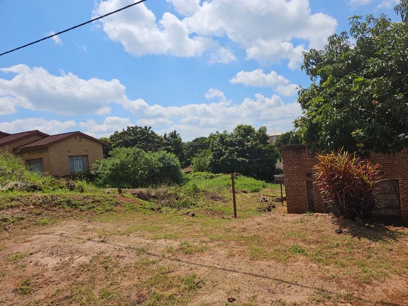 A view of an undeveloped residential plot featuring mature trees and adjacent brick structures, offering potential for future landscaping or construction.