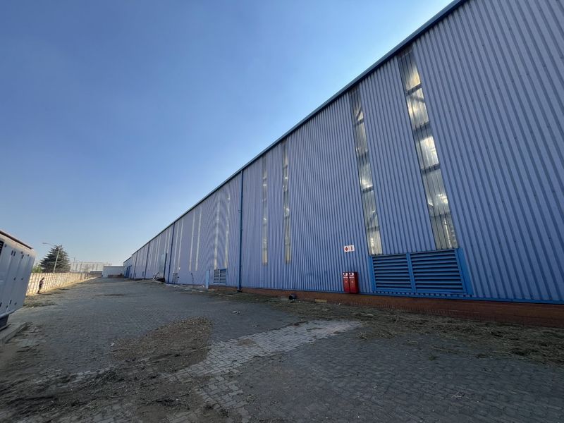 Exterior view of an industrial warehouse featuring corrugated metal cladding, vertical windows, louvered ventilation panels, and a paved yard area.
