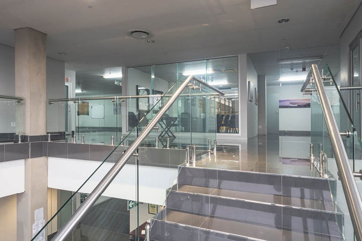 Modern industrial office hallway featuring a glass balustrade staircase, polished tiled flooring, and glass-partitioned office spaces with recessed lighting.
