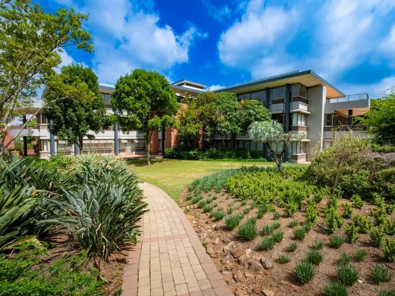 A well-maintained commercial building exterior featuring a paved garden walkway, landscaped grounds, and multi-level office facades with balconies.