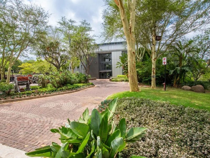 A well-maintained commercial property entrance featuring a paved driveway, lush landscaped gardens, and mature trees leading to a modern building facade.