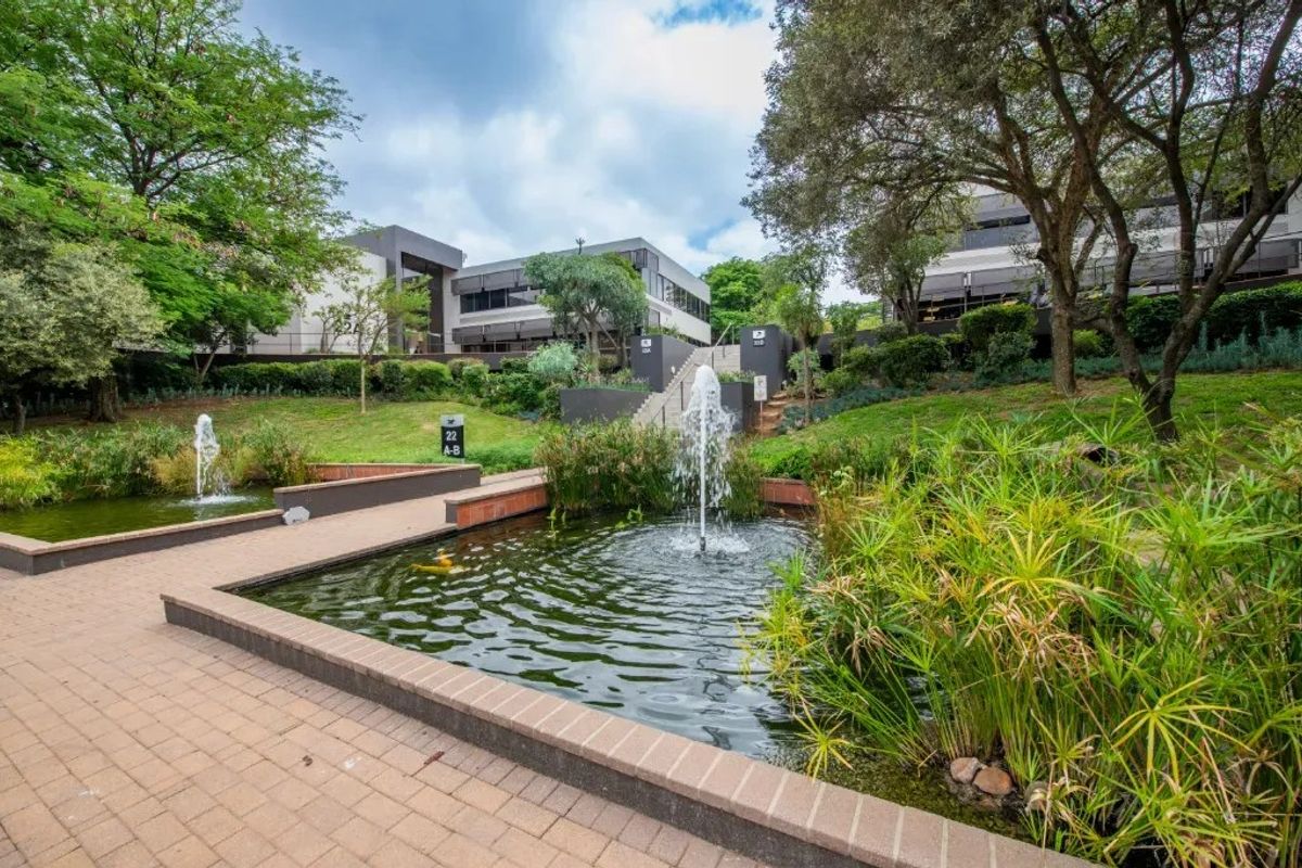 A well-maintained commercial office park exterior featuring landscaped gardens, brick-paved walkways, and decorative water fountains leading to the building entrance.