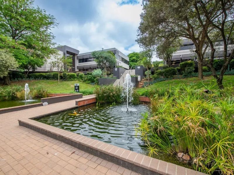 A well-maintained commercial office park exterior featuring landscaped gardens, brick-paved walkways, and decorative water fountains leading to the building entrance.