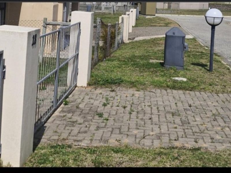 Front entrance area of the townhouse featuring a paved driveway, metal gate, and perimeter fencing with some maintenance required for paving and landscaping.