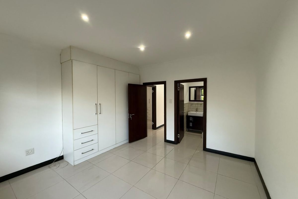 A spacious, empty bedroom featuring neutral tiled flooring, built-in cupboards, recessed downlighting, and direct access to an en-suite bathroom.