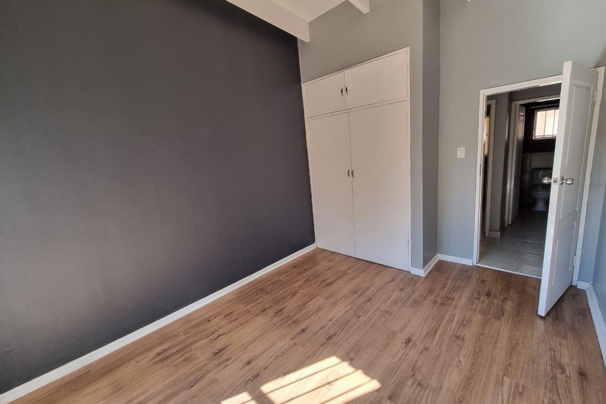A bright, empty bedroom featuring laminate flooring, a built-in wardrobe, exposed ceiling beams, and an en-suite bathroom entrance.