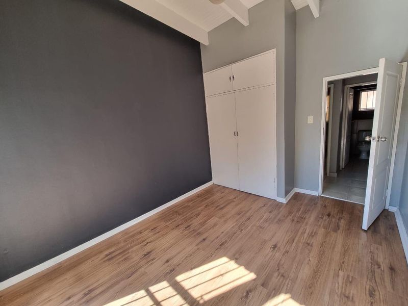 A bright, empty bedroom featuring laminate flooring, a built-in wardrobe, exposed ceiling beams, and an en-suite bathroom entrance.
