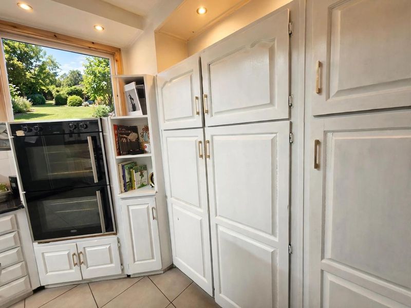 A kitchen area featuring built-in double ovens, integrated white cabinetry with gold hardware, and tiled flooring, with a view of the garden.
