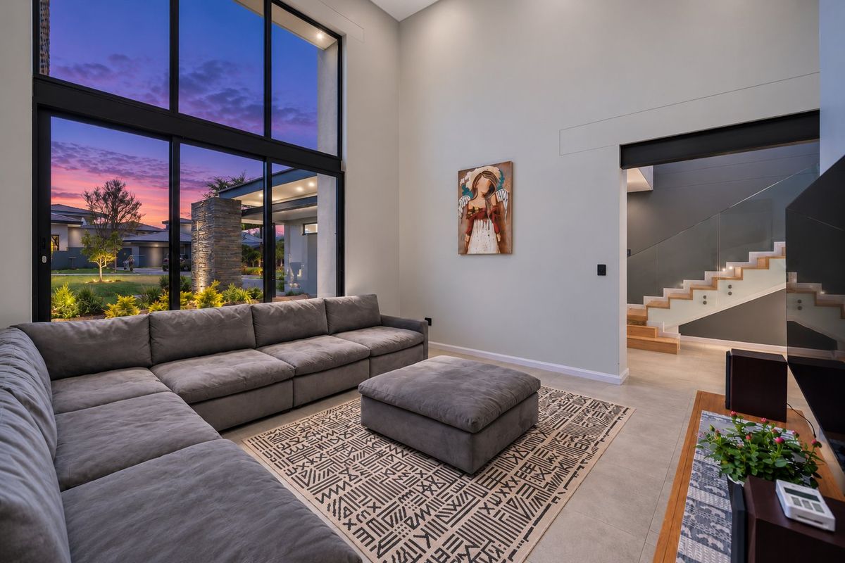 Spacious modern living room featuring double-height floor-to-ceiling windows, tiled flooring, and an elegant staircase with a glass balustrade.