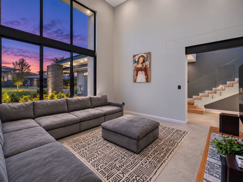 Spacious modern living room featuring double-height floor-to-ceiling windows, tiled flooring, and an elegant staircase with a glass balustrade.