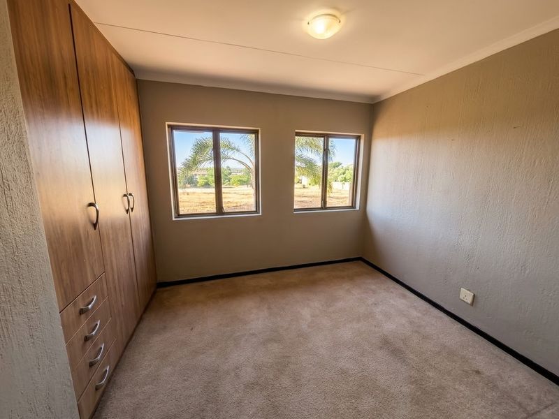 A clean, empty bedroom featuring built-in wooden cupboards, neutral wall finishes, carpeted flooring, and dual windows providing natural light.