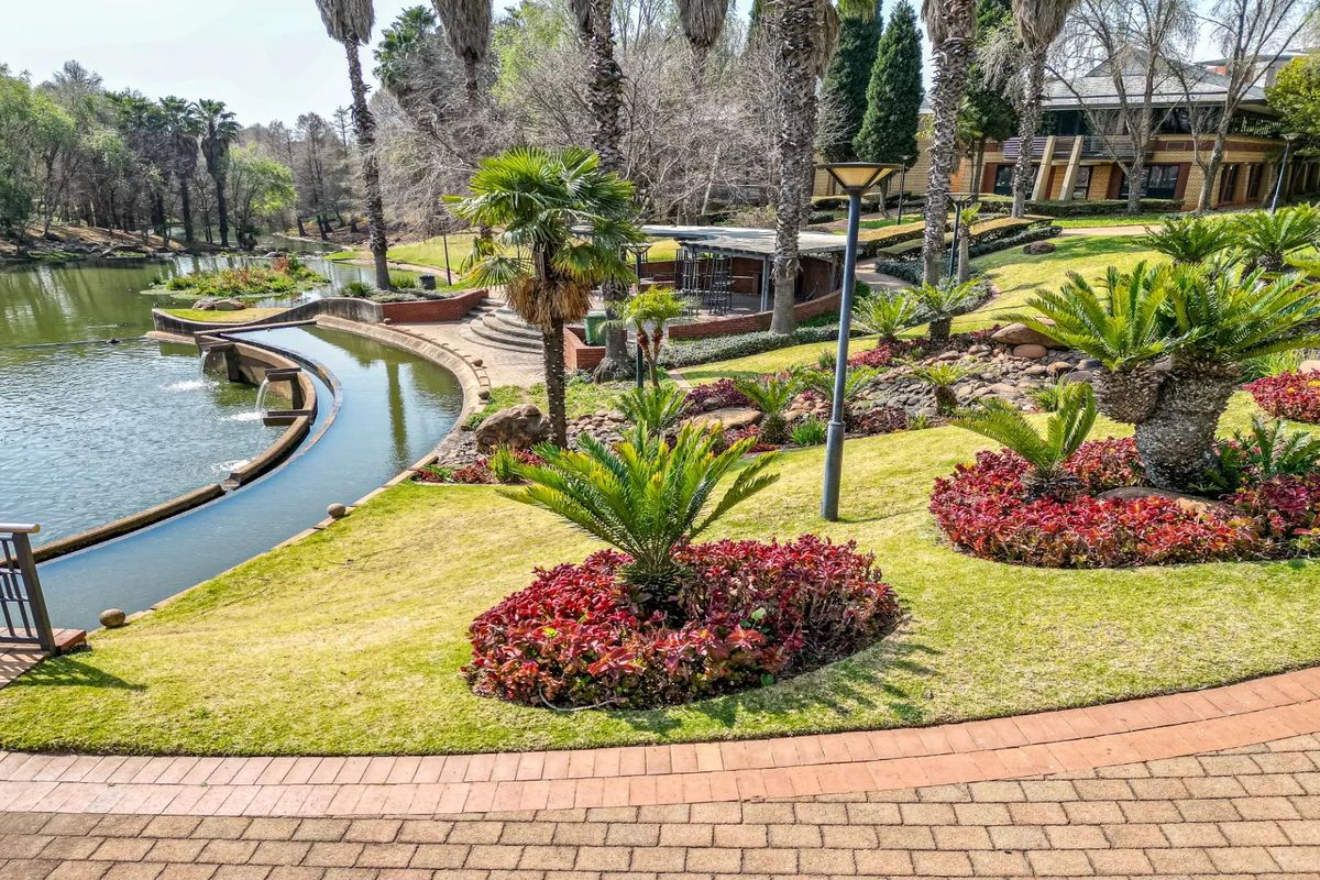 A beautifully landscaped commercial garden featuring manicured lawns, vibrant flower beds, mature palm trees, and a scenic water feature with paved walkways.