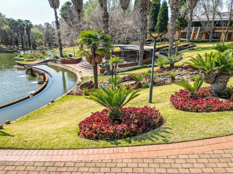 A beautifully landscaped commercial garden featuring manicured lawns, vibrant flower beds, mature palm trees, and a scenic water feature with paved walkways.
