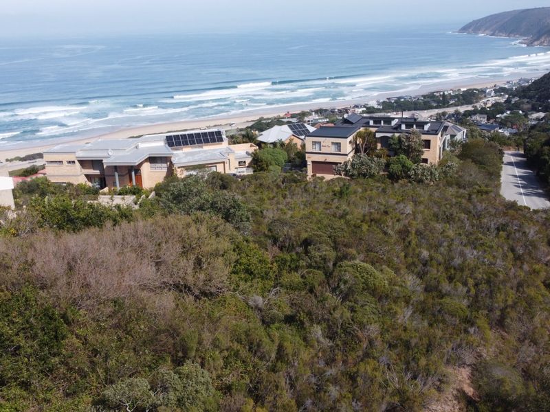 An elevated aerial view of a vacant residential plot featuring dense coastal vegetation, sloping topography, and proximity to the coastline and established residential properties.