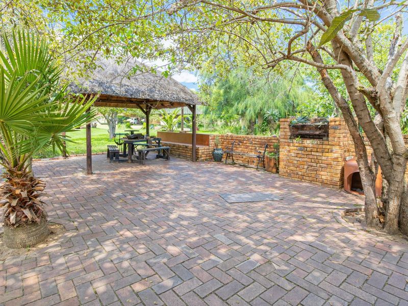 Outdoor entertainment area featuring a paved patio, a thatched gazebo, and a built-in brick braai, set within a garden environment.