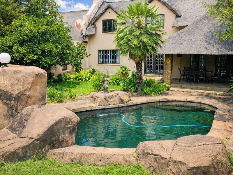 A landscaped backyard featuring a rock-edged swimming pool, a covered patio area, and a multi-story house with a traditional thatched roof.