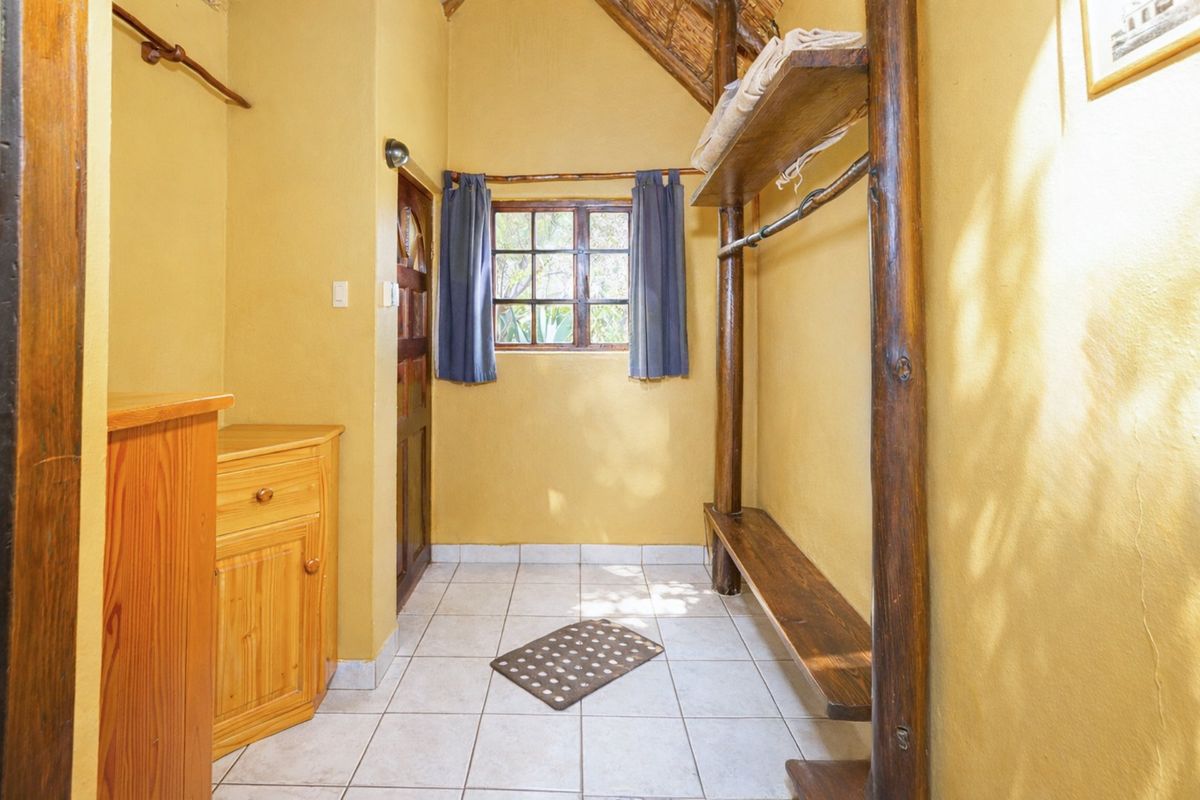 A rustic bedroom featuring a thatched roof, timber structural poles, built-in wooden shelving, and tiled flooring with a window providing natural light.