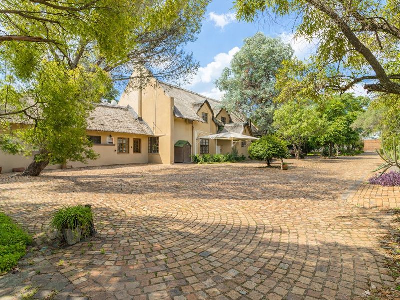 A charming property featuring a traditional thatched roof, stucco exterior, and an expansive, well-maintained cobblestone driveway surrounded by mature trees.