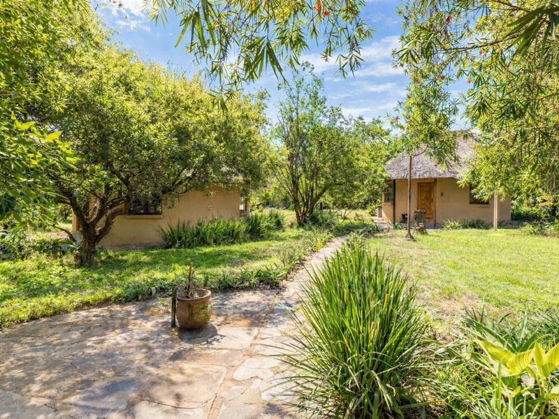 A lush, landscaped garden featuring a stone paved pathway leading between two rustic structures with thatched roofs.