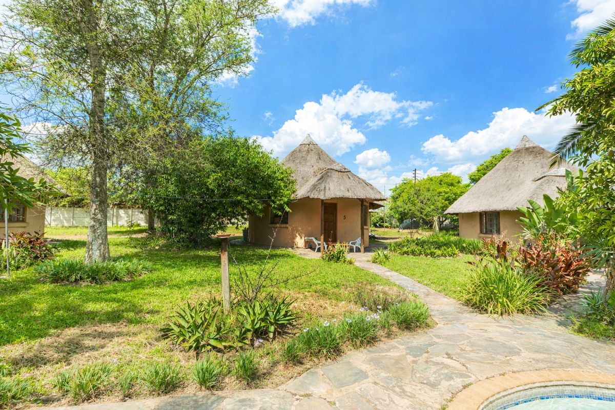 A view of traditional thatched-roof cottages set within a well-maintained garden, featuring stone pathways and access to a swimming pool area.