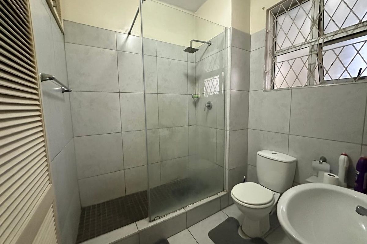 A functional bathroom featuring a glass-enclosed shower with a rain shower head, a pedestal basin, and a toilet, finished with neutral ceramic tiling.