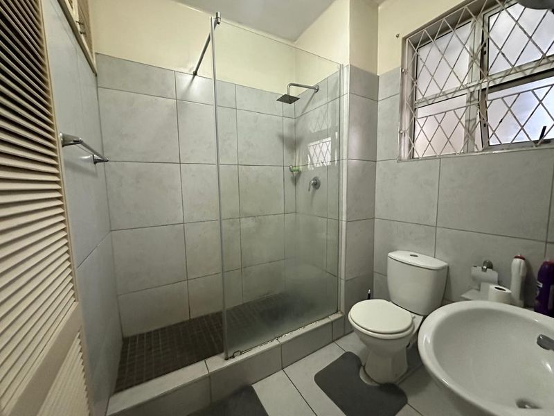 A functional bathroom featuring a glass-enclosed shower with a rain shower head, a pedestal basin, and a toilet, finished with neutral ceramic tiling.