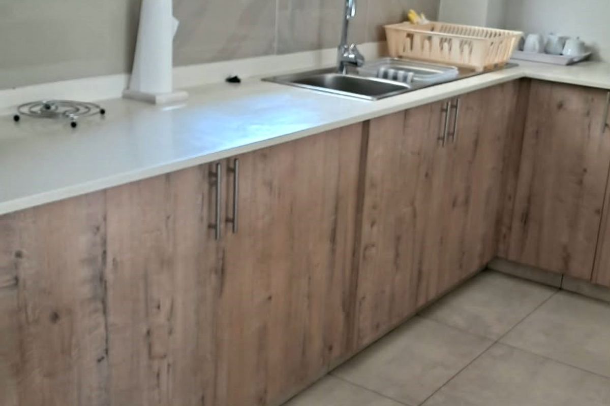 A functional kitchen featuring light wood-look laminate cabinetry, stone countertops, a stainless steel sink, and neutral floor tiles.