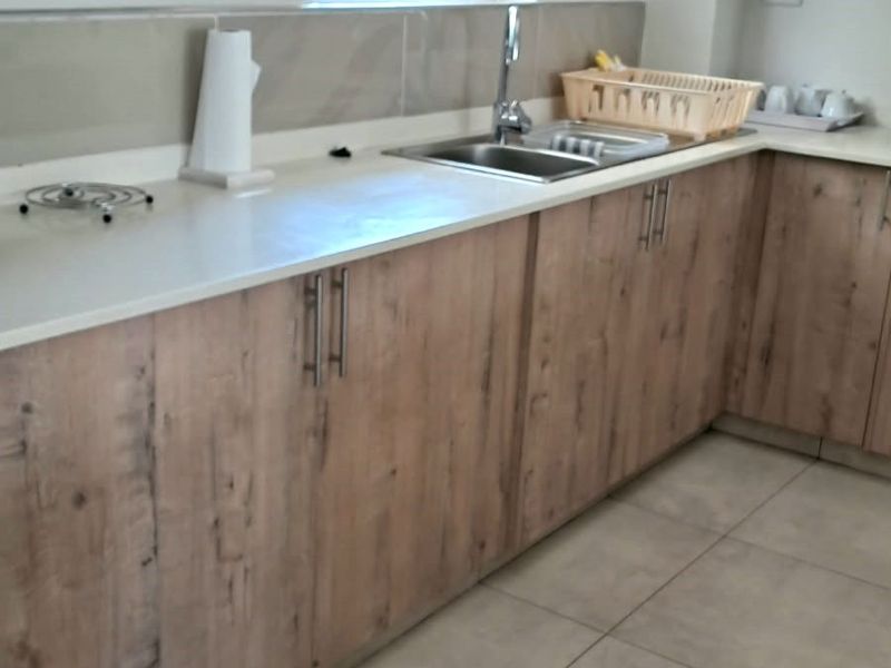 A functional kitchen featuring light wood-look laminate cabinetry, stone countertops, a stainless steel sink, and neutral floor tiles.