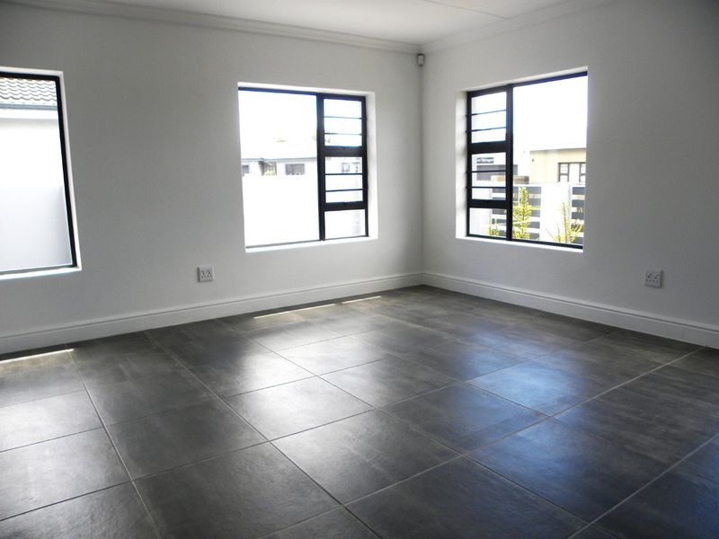 A bright, empty living area featuring modern dark grey floor tiles, crisp white walls, and large aluminium-framed windows providing ample natural light.