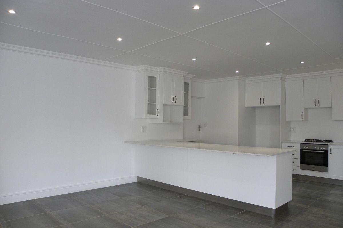 A modern, clean-lined kitchen featuring white cabinetry, stone countertops, a breakfast bar, and integrated stainless steel oven and hob on tiled flooring.