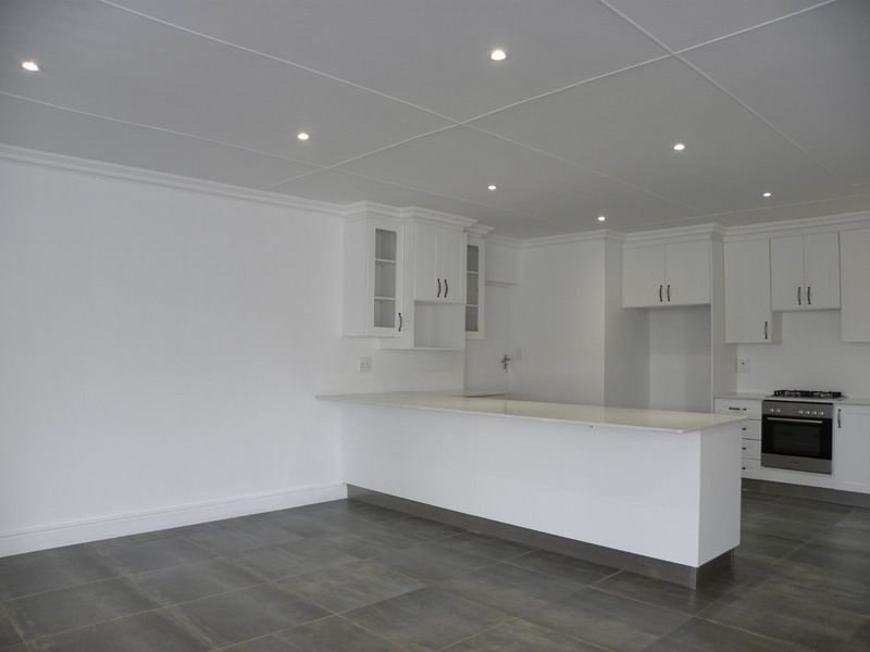 A modern, clean-lined kitchen featuring white cabinetry, stone countertops, a breakfast bar, and integrated stainless steel oven and hob on tiled flooring.
