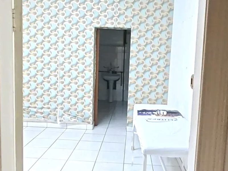 A small commercial bathroom space featuring a pedestal sink, white ceramic floor tiles, and dated patterned wallpaper in need of refurbishment.
