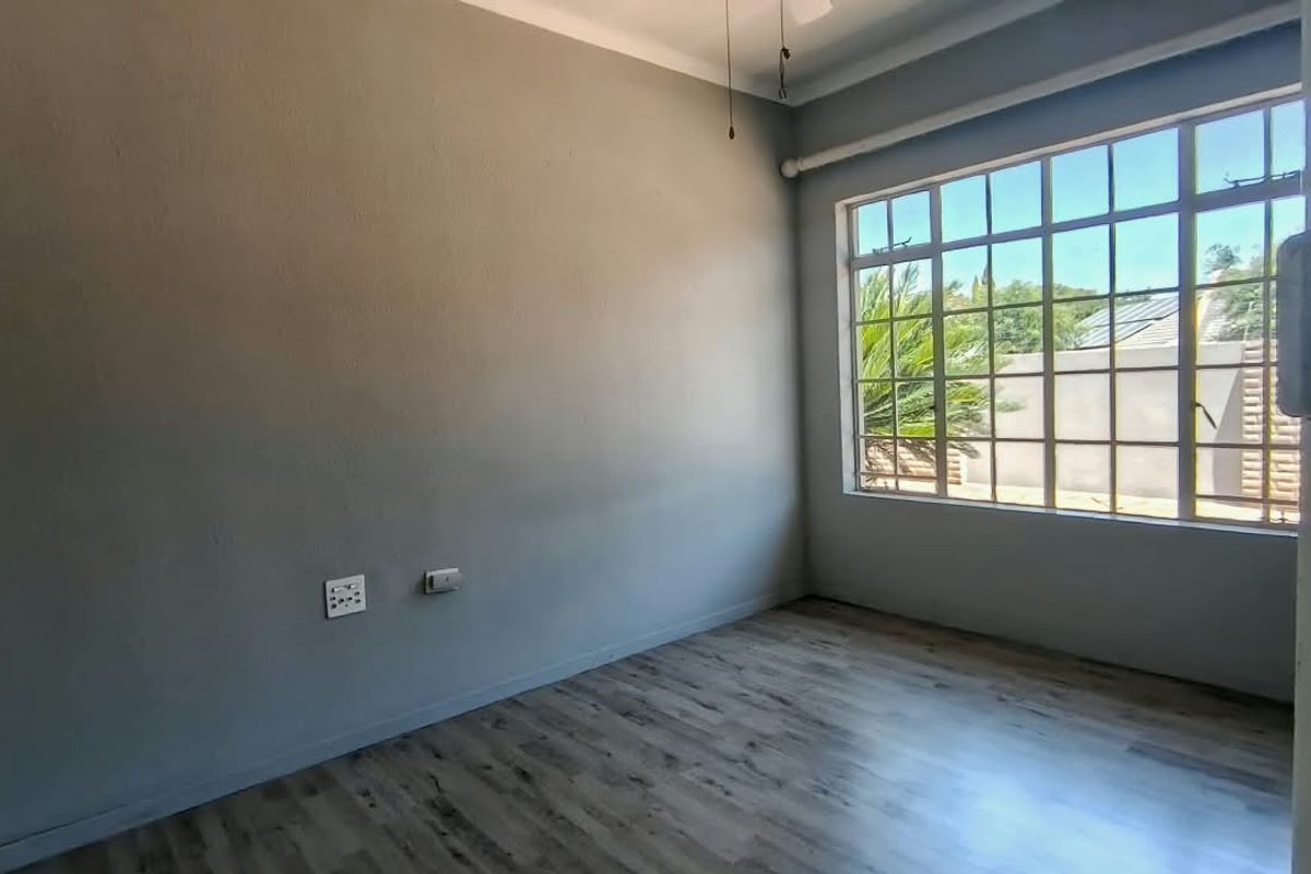 A bright, empty bedroom featuring modern laminate flooring, neutral grey walls, and a large grid-pattern window providing natural light.
