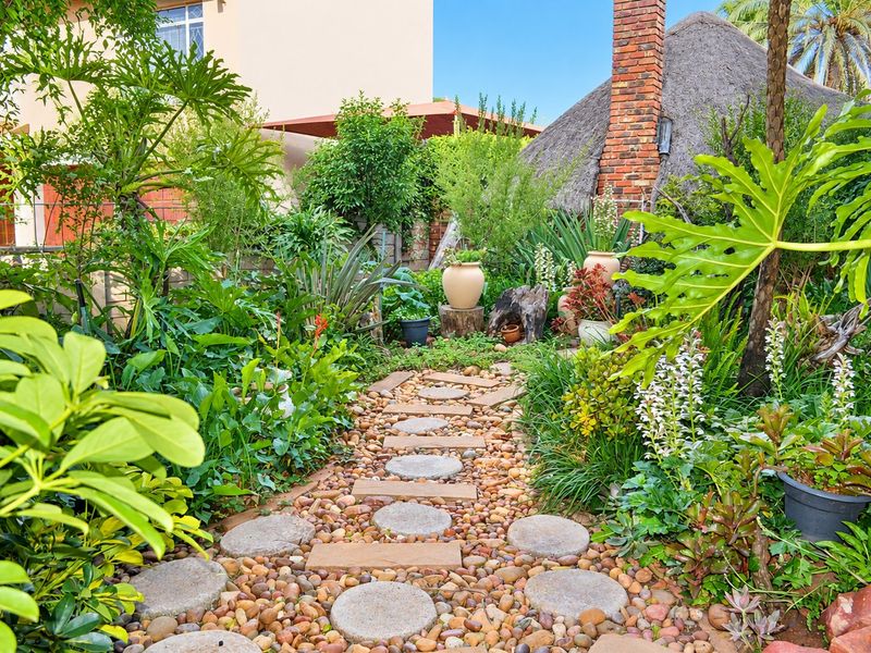 A lush, well-maintained garden featuring a stone pathway, pebble landscaping, and a rustic brick chimney with a thatched roof structure in the background.