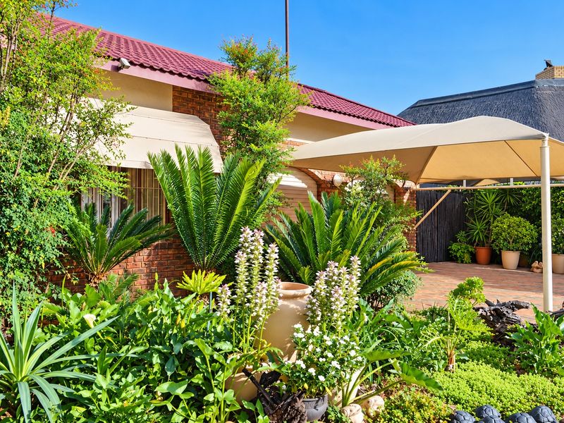 A well-maintained rear garden area featuring a paved patio with a shade awning, established landscaping, and a brick-faced house exterior.