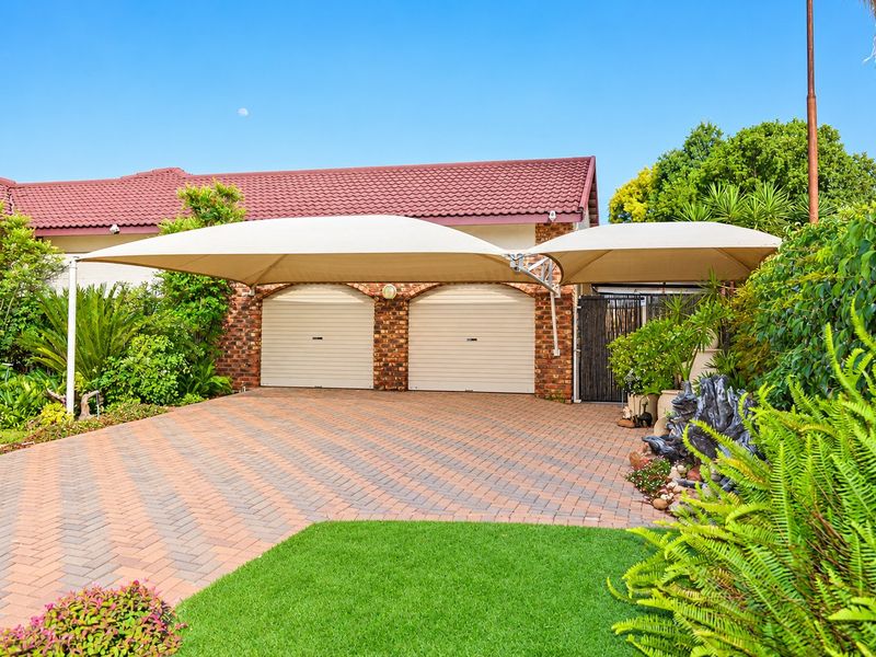 A well-maintained double garage featuring brick exterior, tiled roof, and a paved driveway with an integrated shade sail carport.