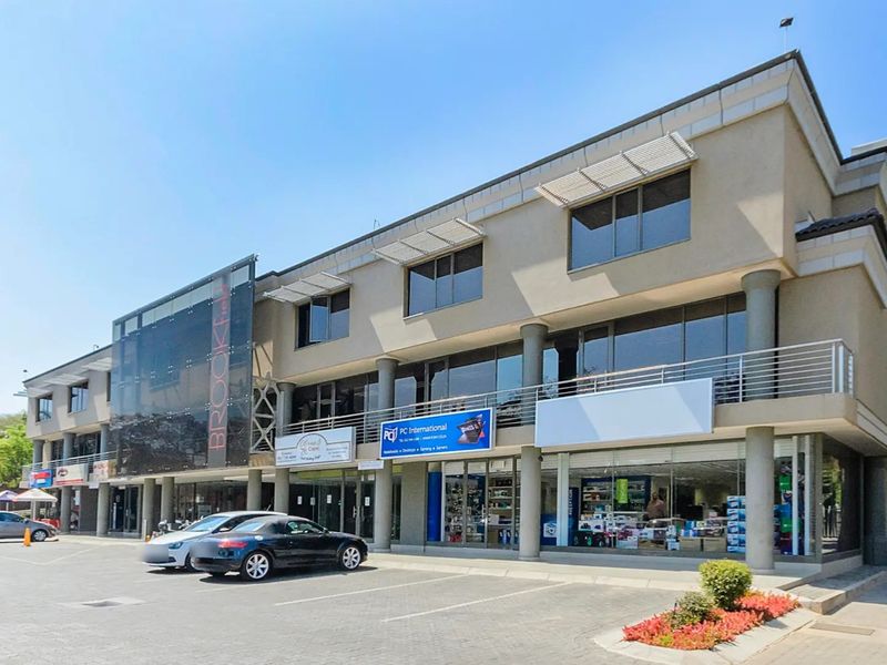 A modern multi-story commercial building featuring glass storefronts, concrete structural columns, and an integrated paved parking area for customer access.