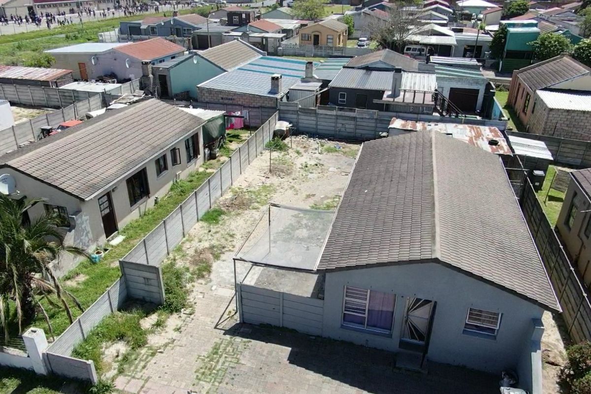 Aerial view of a residential property featuring a tiled roof house, paved driveway, and a large, unmaintained backyard enclosed by precast concrete walls.