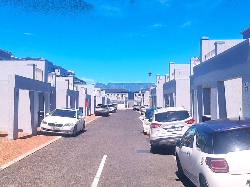 A view of a residential street within a modern housing complex, featuring uniform building facades, paved driveways, and garage access.