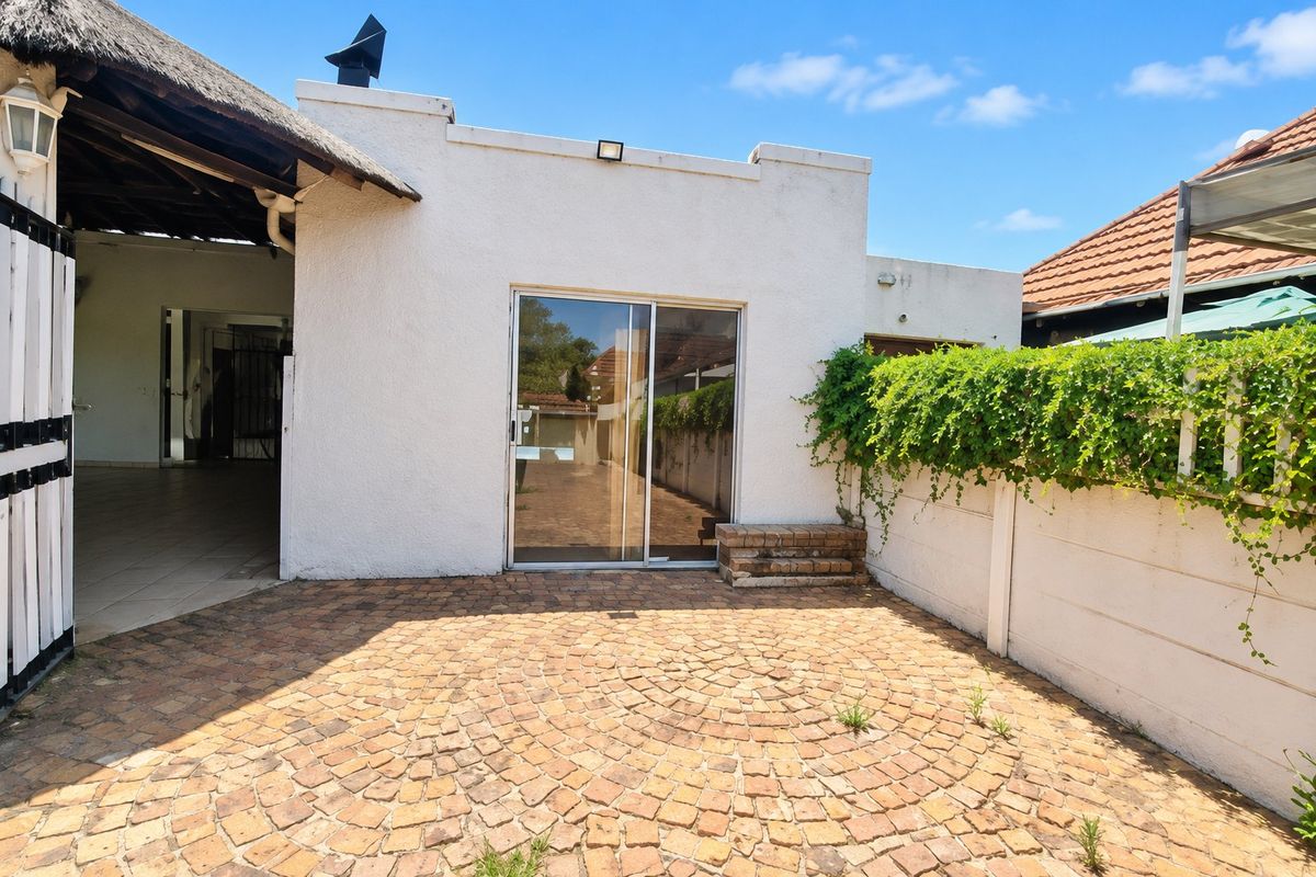 Rear exterior view featuring a brick-paved patio, sliding glass door access, and a thatched roof section, enclosed by a boundary wall with climbing greenery.