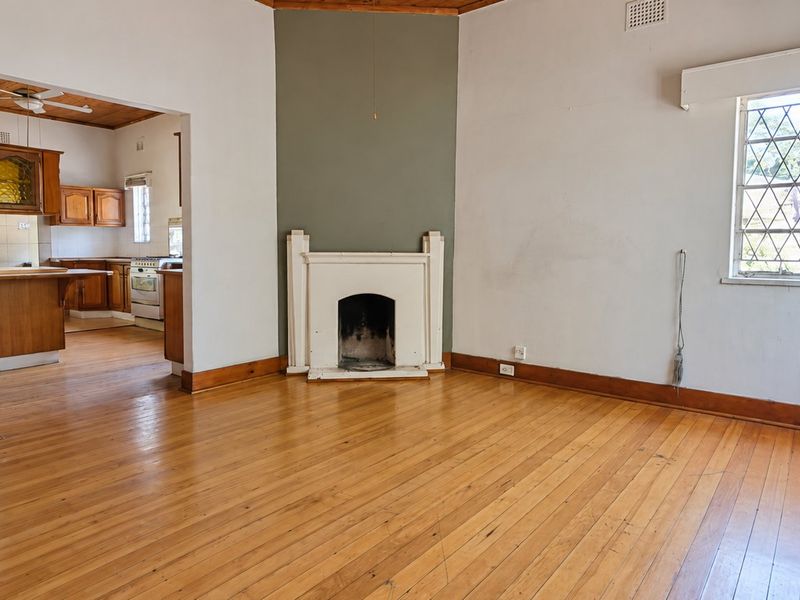 Spacious living room featuring original timber flooring, a central fireplace, and leaded glass windows, with an open-plan flow into the adjacent kitchen area.