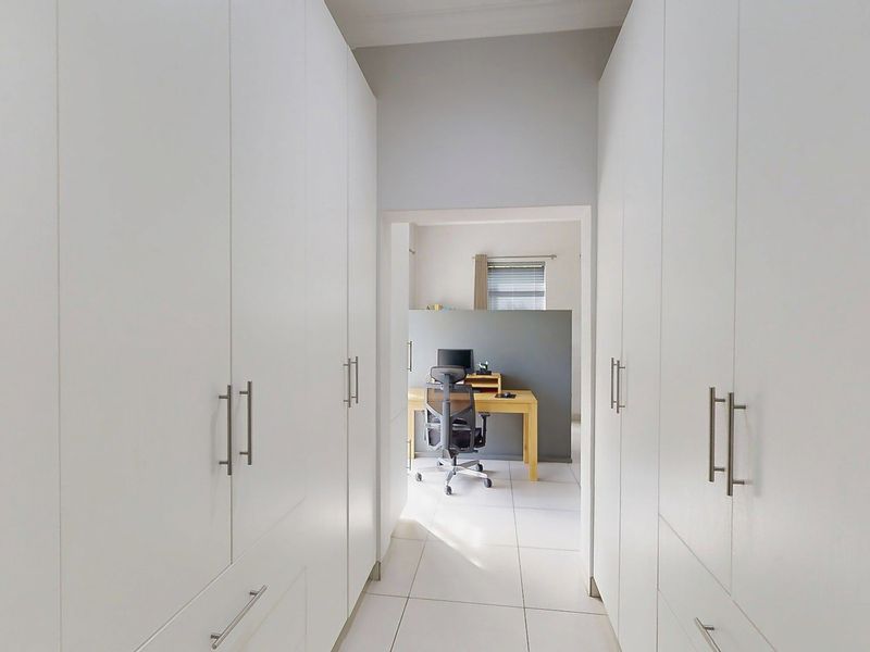 A bright hallway featuring extensive floor-to-ceiling built-in cupboards and clean tiled flooring, leading through to an adjacent office space.