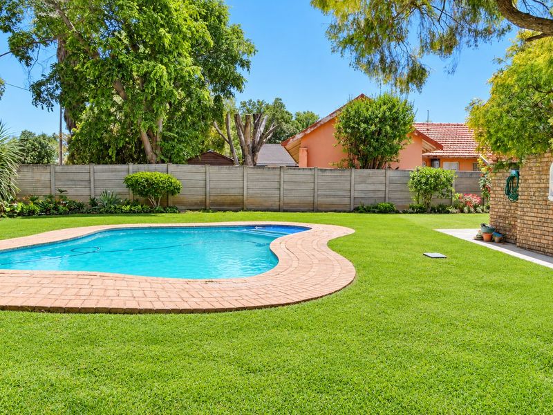 A well-maintained backyard featuring a kidney-shaped swimming pool with brick coping, surrounded by a lush green lawn and secure boundary walling.