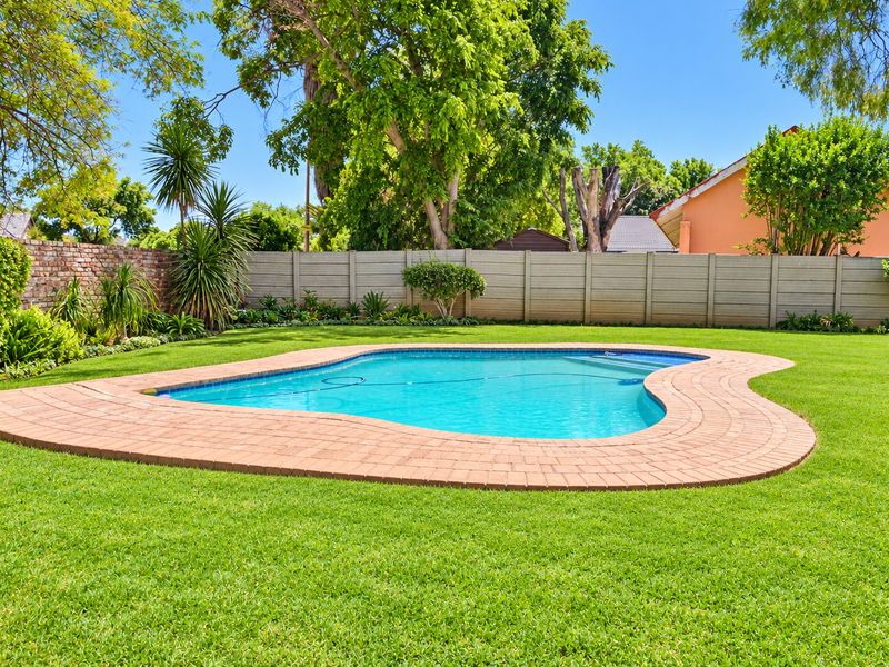 A well-maintained backyard featuring a kidney-shaped swimming pool with brick coping, surrounded by a lush green lawn and secure perimeter walling.