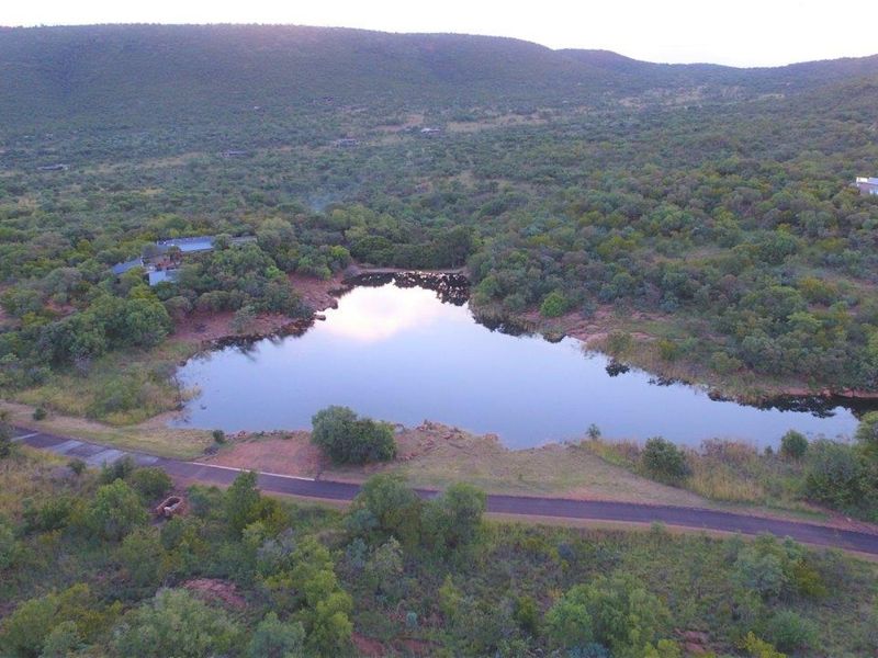 Aerial view of a vacant residential plot featuring a natural dam, established indigenous vegetation, and paved road access within a scenic landscape.