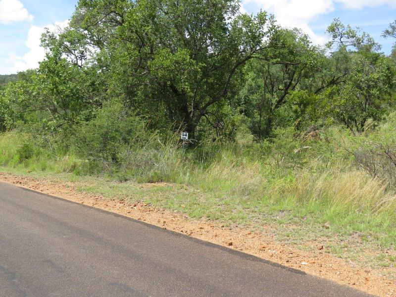 A view of the vacant residential land from the adjacent asphalt road, featuring natural bushveld vegetation and a property boundary marker.